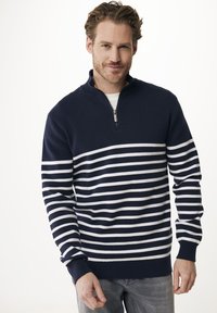 Uomo con capelli e barba castano chiaro, che indossa un maglione a strisce blu navy e bianche con zip a un quarto e jeans grigi, in piedi contro uno sfondo bianco.