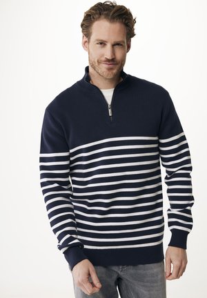 Man met lichtbruin haar en baard, gekleed in een marineblauwe en witte gestreepte sweater met kwartrits en grijze jeans, staand tegen een witte achtergrond.