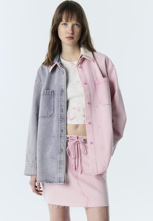 Giovane donna che indossa una giacca di jeans bicolore, un top corto bianco e una gonna di jeans rosa con coulisse in vita su sfondo neutro.