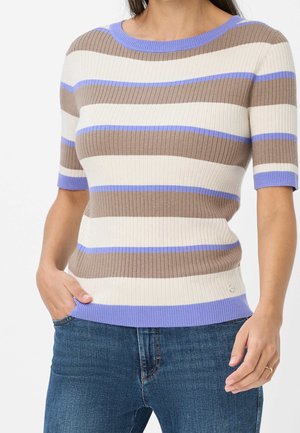Kobieta ubrana w prążkowany sweter z krótkim rękawem w poziome beżowe, białe i jasnofioletowe paski oraz niebieskie dżinsy, stojąca z jedną ręką w kieszeni.