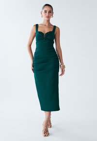 Green fitted dress with a sweetheart neckline, ruched texture, wide straps, and a center detail. Below-knee length, paired with strappy sandals.