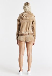 Fløjlsagtig tan hættetrøje med broderet logo, kombineret med matchende shorts. Har snøre ved hætten og blød tekstur. Hvide sneakers fuldender looket.