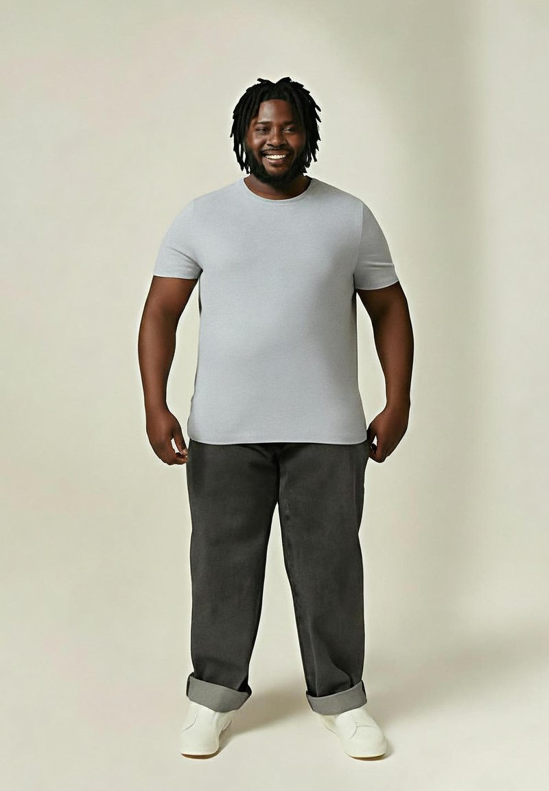 Homme souriant avec des dreadlocks de taille moyenne portant un t-shirt gris clair, un pantalon foncé retroussé et des baskets blanches, se tenant sur un fond neutre.