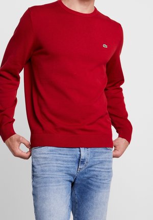 Homme portant un pull rouge uni à manches longues avec un petit logo vert sur la poitrine et un jean bleu clair, ajustant l'ourlet du pull.