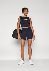 Svart atletisk crop-top med "NEBBIA" tekst, høyhælte svarte shorts og hvite sneakers. Modellen holder en svart treningsbag. Glatt stofftekstur.