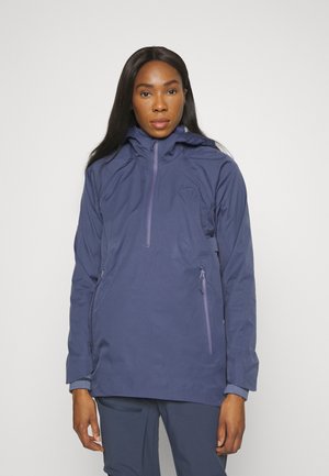 Γυναίκα φορώντας μια ναυτικά μπλε μπουφάν με κουκούλα και δύο τσέπες με φερμουάρ, καθώς και ταιριαστούς ναυτικούς παντελόνες, που στέκεται μπροστά σε απλή ανοιχτή φόντο.