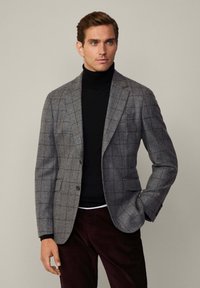 Blazer grigio a quadri con accenti rossi, due bottoni e tasche a patta, indossato su un dolcevita nero e pantaloni bordeaux. Tessuto morbido.