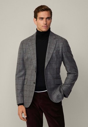 Grauer Karoblazer mit roten Akzenten, zwei Knöpfen und Klappentaschen, getragen über einem schwarzen Rollkragenpullover und burgunderfarbenen Hosen. Glatte Textur.