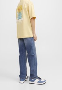 Gul bomullströja med blommig grafik på baksidan, tillsammans med ljusblå denimjeans och blåvita sneakers. Minimalistisk design.