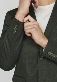 Dark green blazer with a textured fabric, featuring three button accents on the cuff and a notch lapel. Worn over a white shirt.