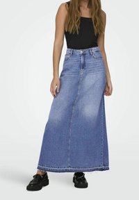 Kvinna som bär en lång blå denimkjol med fläkt nederkant, svart ärmlös topp och svarta loafers, stående mot en enfärgad bakgrund.