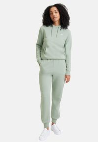 Sudadera verde clara y joggers a juego, fabricados en un tejido suave, con un bolsillo frontal, capucha con cordón y puños acanalados. Zapatillas blancas.