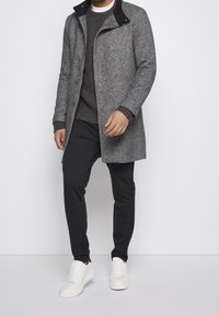 Grauer Wollmischmantel mit schwarzem Kragen, kombiniert mit einem strukturierten dunklen Pullover und schwarz gestreiften Hosen, abgerundet mit weißen Sneakern.