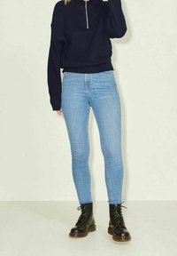 Henkilö, joka on pukeutunut vaaleansinisiin kapeihin farkkuihin, mustiin nauhakenkiin ja tummansiniseen vetoketjupuseroon, jossa on pitkät hihat.