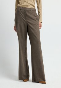 Vrouw in bruin hoge taille brede broek en een beige lange-mouwen knoopshirt, staand tegen een effen witte achtergrond.