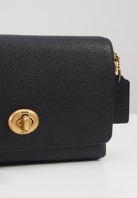 Sac bandoulière en cuir noir avec une finition texturée, des ferrures dorées et une fermeture à fermeture tournante signée. Comprend un détail de tag suspendu.