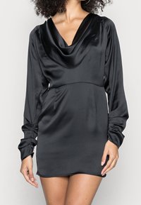 Musta satiininen minimekko, jossa on kaarroke-kaula-aukko, pitkät istuvat hihat ja sivutaskut. Pehmeä pinta, joka heijastaa valoa, ja muotoiltu siluetti.