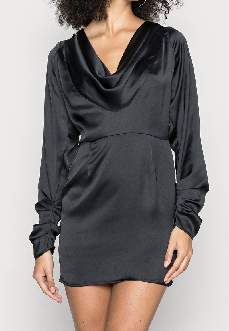 Musta satiininen minimekko, jossa on kaarroke-kaula-aukko, pitkät istuvat hihat ja sivutaskut. Pehmeä pinta, joka heijastaa valoa, ja muotoiltu siluetti.