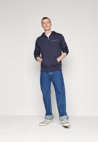 Felpa con zip color navy, dotata di tasche frontali e logo, abbinata a jeans dritti blu e scarpe grigie. Il modello si trova in posizione frontale.