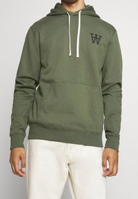 Sudadera verde de algodón con un bolsillo delantero y cordones. Presenta un pequeño logotipo negro en el pecho y puños acanalados.