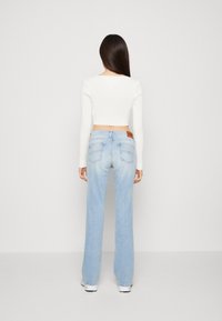 Donna in piedi, di spalle, che indossa un top cropped a maniche lunghe bianco, jeans a vita alta di un blu chiaro e sneakers bianche su uno sfondo neutro.