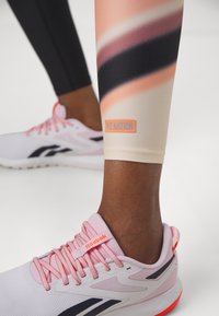 P.E Nation legginssit, joissa on vaaleneva design persikanvärisestä, vaaleanpunaisesta ja mustasta, yhdistettynä valkoisiin ja vaaleanpunaisiin Reebok-tennareihin, joissa on yksityiskohtaiset nauhat.