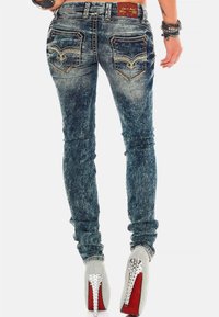 Jeans blu skinny in denim con texture sbiadita, tasche posteriori decorative e dettagli di cucitura, abbinati a scarpe col tacco alto con accenti argentati.