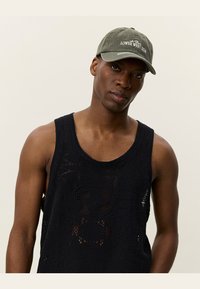 Oliven grønn baseballcaps med hvit brodert tekst, kombinert med en marineblå ermeløs strikket tanktop med et åpent, teksturert mønster.