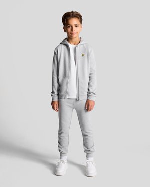 Graue Zip-Hoodie- und Jogginghosen-Kombination aus weichem Stoff. Darunter trägt die Person ein weißes T-Shirt und weiße Sneaker. Kleines gelbes Logo auf dem Hoodie.