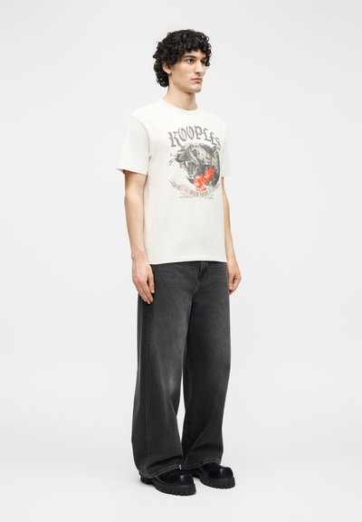 Νέος άντρας με σγουρά μαλλιά, φορώντας μια εκρού μέγεθος μπλούζα με το γράφημα μιας βρυχώμενης πάνθηρας και φαρδιά μαύρα τζιν παντελόνια, στέκεται μπροστά σε ένα απλό φόντο.