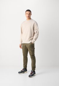 Beżowa bluza z okrągłym dekoltem, oliwkowe spodnie cargo z kieszeniami oraz czarne sneakersy z granatowymi i bordowymi akcentami. Gładkie faktury.