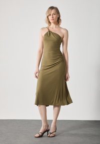 Robe midi olive vert, à une épaule, avec des détails froncés sur le côté, texture côtelée et silhouette ajustée. Portée avec des talons noirs à lanières.