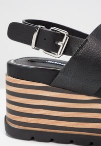 Sandale compensée en cuir noir avec bride à boucle argentée et semelle plateforme à motif bois superposée avec des rayures noires.