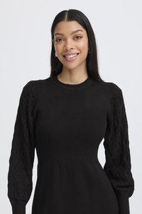 Abito nero con corpetto a coste e maniche balloon a trecce in maglia testurizzata. Tessuto liscio con una sottile lucentezza e silhouette aderente.