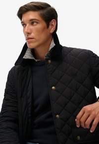 Czarna pikowana kurtka z kordurowym kołnierzem i w kratkę podszewką, z ozdobnymi guzikami i teksturowanym ciemnoniebieskim swetrem pod spodem.