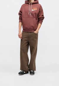 Personne portant un sweat à capuche bordeaux avec des cordons blancs, un pantalon large marron, des baskets noires et blanches, debout avec une main dans la poche.