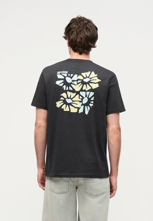 Άνδρας που φοράει μαύρο μπλουζάκι με μπλε και κίτρινο αφηρημένο σχέδιο λουλουδιού στην πλάτη, συνδυασμένο με ανοιχτό γκρι τζιν σορτς.