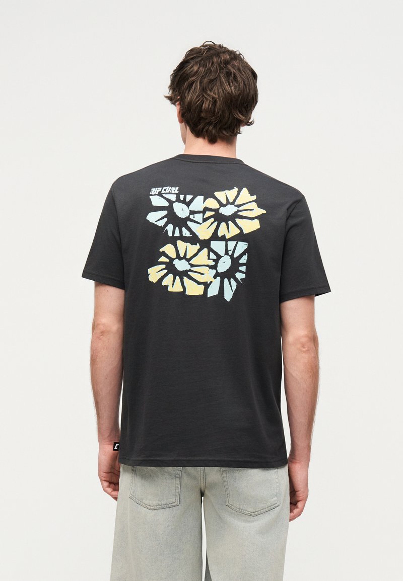 Άνδρας που φοράει μαύρο μπλουζάκι με μπλε και κίτρινο αφηρημένο σχέδιο λουλουδιού στην πλάτη, συνδυασμένο με ανοιχτό γκρι τζιν σορτς.