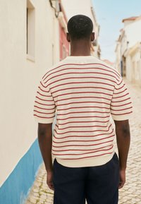 Creme strikket sweater med røde striber, korte ærmer, rund halsudskæring og ribbede manchetter, parret med mørke bukser. Dæmpet gadebaggrund.