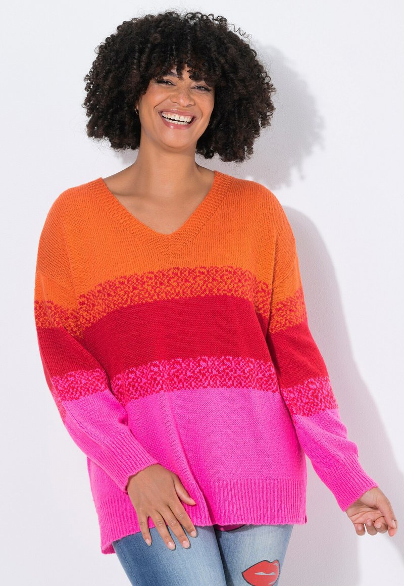 Donna con capelli ricci che sorride, indossando un maglione a maglia multicolore con strisce arancioni, rosse e rosa, e jeans blu con una toppa a forma di labbra.