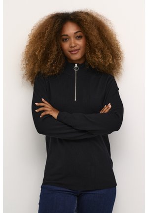 Sort strikket sweater med høj krave og sølv lynlåsdetalje. Har ribbet tekstur foran og lange ærmer. Bæres sammen med blå jeans.