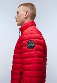 Rød pufferjakke med høj krave. Har et rundt logo-patch på venstre ærme, horisontale baffelsyninger og lynlås-lommer.