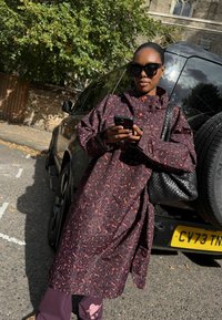 Naine, kes kannab tumeda mustriga pikkade mantlit ja päikeseprille, seisab musta maasturi kõrval, kasutades väljas varjus nutitelefoni.