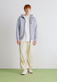 Felpa grigia chiara con zip e tasche, indossata sopra una maglietta bianca, abbinata a pantaloni cargo beige e sneakers bianche su un pavimento verde.