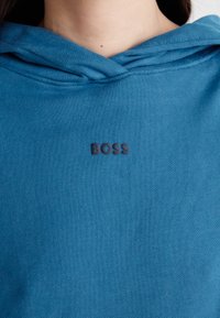 Felpa blu con cappuccio e cordino, dal taglio rilassato, con un logo "BOSS" ricamato in nero sul petto. Texture liscia.