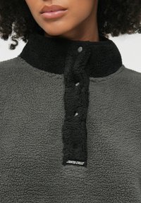 Φλιντζάνι πούλοβερ με μαύρο κολάρο και κουμπότρυπα, με στιβαρό γκρι σώμα και ετικέτα «Santa Cruz» στο πλάι.