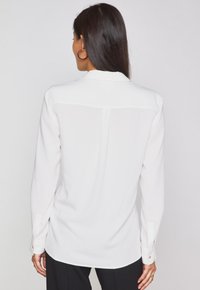 Blusa bianca a maniche lunghe con polsini abbottonati, texture liscia, colletto classico e leggero lucido. Il retro presenta un dettaglio di cucitura centrale.