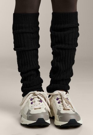 Fekete bordás lábszármelegítők ráncolt textúrával, sötét leggings felett viselve, világosszürke és fehér sportcipőkkel párosítva.