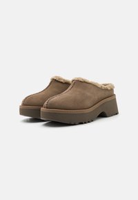 Zoccoli in suede marrone con una morbida fodera beige, una suola platform spessa e cuciture in contrasto lungo il bordo. Grip texturizzato sulla suola.