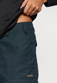 Mørk teal G-1000 stoffbukse med glatt tekstur, med en frontlomme og fremhevede sømmer. Gullfarget "G-1000"-merke synlig.